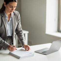 Consultant indépendant consultant et comparant des documents de mission devant un bureau sobre, avec un ordinateur portable, dans un bureau moderne éclairé par la lumière du jour.
