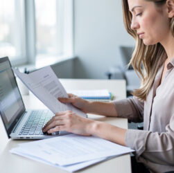 Une femme travaille sur un ordinateur portable dans un bureau moderne, consulte des documents imprimés et organise des dossiers, dans une ambiance professionnelle lumineuse.